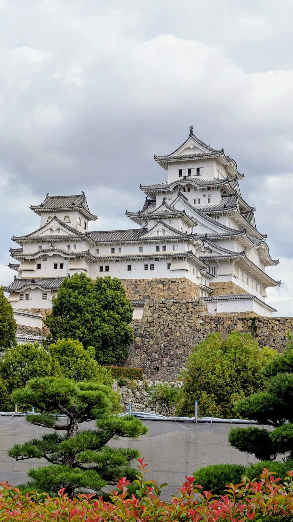 Il castello bianco di Himeji su mura di pietra, con pini e fiori davanti