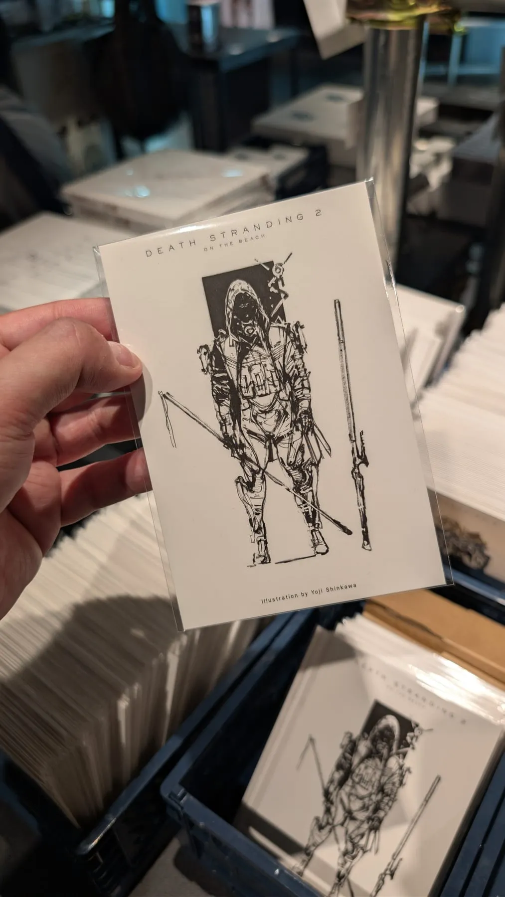 Mano che tiene una cartolina disegnata di Death Stranding 2 in un negozio